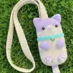 The finished purple and beige crochet cat phone bag with its long strap lying flat on green grass