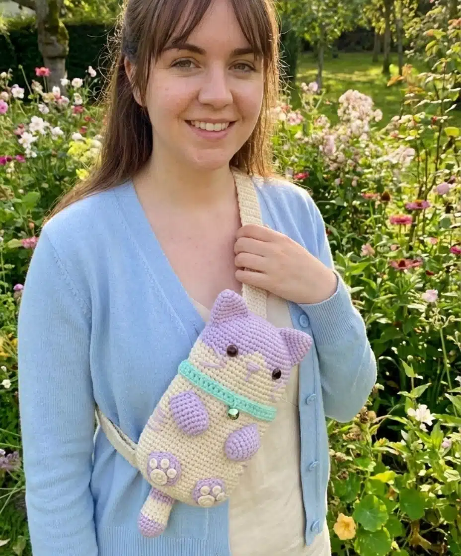 A woman in a blue cardigan wearing the purple and beige crochet cat phone bag as a crossbody bag in a garden setting.
