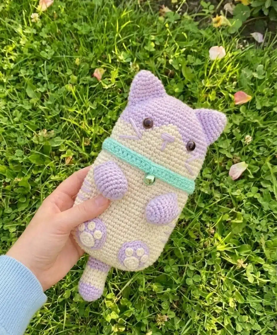 Hand holding a completed purple and beige crochet cat phone bag with a teal collar and bell, resting on green grass.